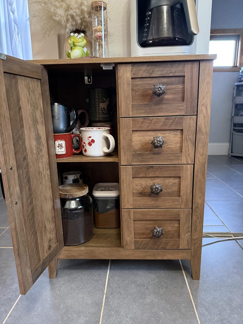 May include: A wooden coffee cabinet with four drawers and a cabinet door. The drawers have decorative floral knobs. The cabinet door is open, revealing mugs, a jar of coffee, and a container. A coffee maker sits on top.