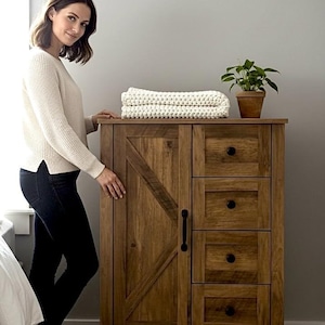 May include: A wooden cabinet with a rustic design. The cabinet has a barn door style on the left and three drawers on the right. A stack of cream-colored blankets and a potted plant sit on top. The cabinet is made of wood and has a dark brown finish.