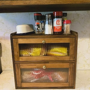 May include: A wooden spice rack with two glass-fronted drawers and a top shelf. The drawers are filled with packets, and the top shelf holds spice jars and a small white container. The wood has a warm, brown stain.