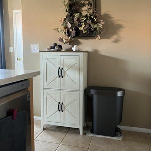 May include: A white cabinet with four doors and black handles. The doors have a decorative X design. A black trash can sits next to the cabinet. A decorative wall piece with greenery is above the cabinet.
