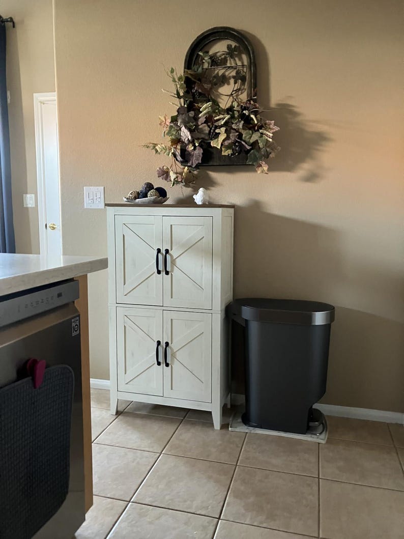 May include: A white cabinet with four doors and black handles. The doors have a decorative X design. A black trash can sits next to the cabinet. A decorative wall piece with greenery is above the cabinet.