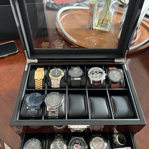 May include: A black wooden watch display case with a glass lid, featuring two tiers of storage compartments. The top tier holds seven wristwatches with various bands, while the bottom drawer contains four additional watches and a silver ring.