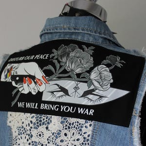 May include: A denim vest with a distressed hem and a large black patch on the back. The patch features a hand holding a knife, flowers, and the text "DISTURB OUR PEACE WE WILL BRING YOU WAR." A white lace panel is sewn on the back.