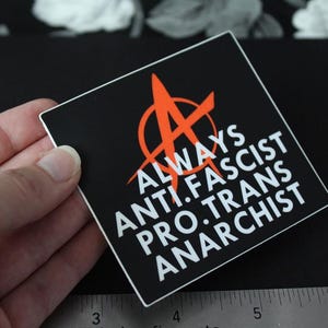 May include: A square black sticker featuring a red anarchist symbol and white text that reads ALWAYS ANTI-FASCIST PRO-TRANS ANARCHIST. The sticker is positioned above a metal ruler showing measurements in inches, with the edge at the 3-inch mark.