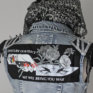 Pode incluir: Um colete de ganga de lavagem clara com um patch preto com uma mão segurando uma faca, flores e o texto "DISTURB OUR PEACE WE WILL BRING YOU WAR". O colete tem detalhes de lantejoulas prateadas, franjas e um capuz com lantejoulas.