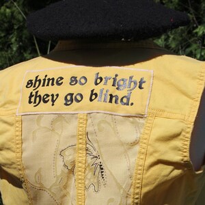 May include: A yellow sleeveless vest featuring a rectangular patch on the upper back with the black embroidered text "shine so bright they go blind." The center panel displays a light yellow fabric with a subtle butterfly and vine pattern.
