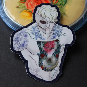 May include: A sticker featuring a stylized illustration of a person with a half-skull face and a floral design. The chest cavity reveals a heart and more floral elements. The sticker has a dark blue border.