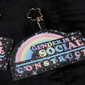 May include: Two keychains with the text "GENDER IS A SOCIAL CONSTRUCT" over a rainbow design. The keychains have a black background with silver glitter and star accents. One keychain has a star-shaped clasp, the other has a flower-shaped clasp.