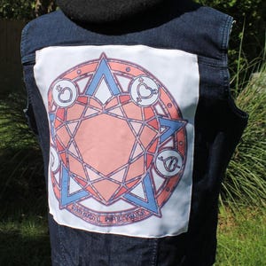 May include: A dark blue denim vest featuring a large white square patch on the back. The patch displays a circular geometric design in shades of pink and blue, incorporating various symbols and the text "MAGICAL SPECTRUM" at the bottom.