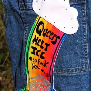 May include: A blue denim garment featuring a white cloud patch with small metallic studs. Below the cloud is a rainbow-colored graphic with the black text: Queers Melt Ice Also Fuck You, The 216, and @goodboymorgan, ending in a silver glitter puddle.