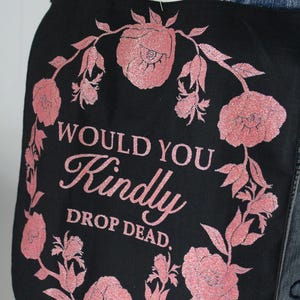 May include: Black tote bag with a pink floral wreath design. The text within the wreath reads "WOULD YOU Kindly DROP DEAD." The flowers and text have a glittery finish. The bag is worn over a denim jacket.