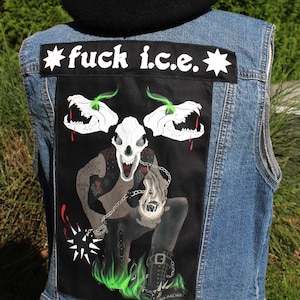 May include: A blue denim vest featuring a large rectangular black patch on the back. The patch displays a graphic of a creature with a skull head, green flames, and chains, with the text "fuck i.c.e." printed in white at the top.