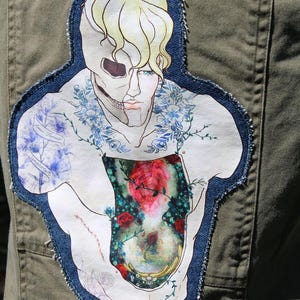 Könnte beinhalten: Eine olivgrüne Jacke mit einem großen, unregelmäßig geformten Stoffaufnäher auf der Rückseite. Der Aufnäher zeigt eine stilisierte Illustration einer Figur mit einem halben Totenkopfgesicht, blondem Haar, blauen Blumendetails und einem mit roten Rosen gefüllten Oberkörper.