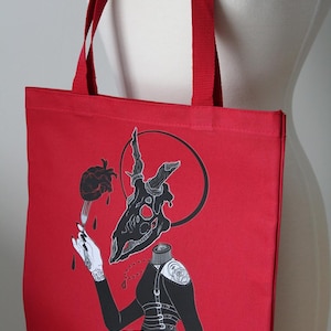 Può includere: Borsa tote rossa con illustrazione in bianco e nero. L'opera presenta un teschio di cervo, un cuore su una forchetta e una figura senza testa con una giacca nera. La borsa ha manici rossi ed è esposta su un manichino.