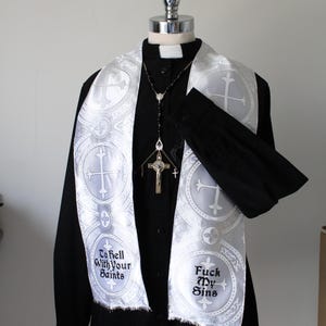 May include: A black long-sleeved shirt with a white clerical collar and a white liturgical stole featuring circular cross patterns. The stole displays the embroidered text "To Hell With Your Saints" and "Fuck My Sins" with a rosary necklace.