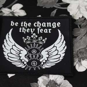 May include: A rectangular black fabric patch featuring white screen-printed graphics. The design includes a pair of wings, a crown, and two stylized eyes with symbols in the pupils. The text "be the change they fear" is printed in white.