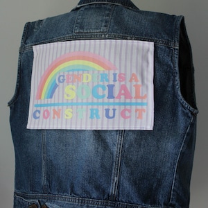 May include: A blue denim vest featuring a patch with the text "GENDER IS A SOCIAL CONSTRUCT" in pastel colours above a rainbow. The vest is a medium wash and has a classic design.
