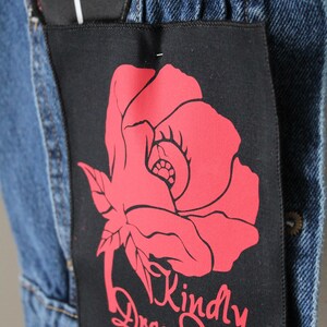 May include: Close-up of a denim jacket with two black patches. One patch has the words "DROP DEAD" in white. The other patch features a red rose graphic and the words "Kindly Drop Dead" in red. The jacket is blue.