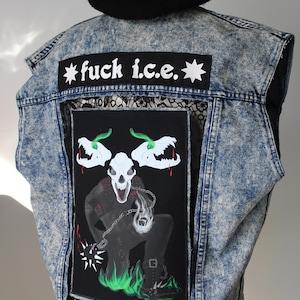Puede incluir: Chaleco vaquero con lavado ácido y boina negra. El chaleco presenta un parche grande con un gráfico de calavera y el texto "fuck i.c.e." en blanco. La parte trasera del chaleco tiene un panel de encaje negro.