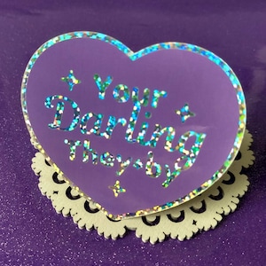 May include: A purple heart-shaped pin with the text "Your Darling They/by" in glittery letters. The pin has a holographic border and is set on a white, scalloped base. The background is a textured purple surface.