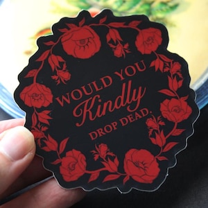 May include: Black sticker with red floral border and text that reads "WOULD YOU Kindly DROP DEAD." The sticker is held by a hand. The sticker is in front of a blurred background.