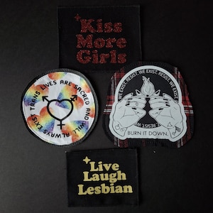 May include: Four fabric patches with text and graphics. One patch reads "Kiss More Girls" in red glitter. Another says "Trans Lives Are Sacred And Will Always Exist" with a rainbow heart symbol. A third patch says "Burn It Down". The last patch says "Live Laugh Lesbian".