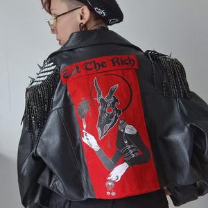 Könnte beinhalten: Schwarze Lederjacke mit einem roten Aufnäher mit einem grafischen Design und dem Text "Eat The Rich". Die Jacke hat Fransen und Spikendetails an den Schultern. Ein schwarzer Barett mit weißen Augendesigns ist ebenfalls sichtbar.