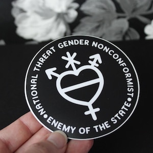 May include: A circular black sticker with white text and a central graphic. The text reads "GENDER NONCONFORMIST" and "NATIONAL THREAT ENEMY OF THE STATE". The central design features a heart shape combined with various gender symbols.