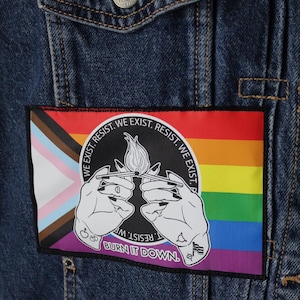 May include: A dark blue denim jacket featuring a rectangular patch with a pride flag background. The patch displays two hands holding a lit match, surrounded by the text "WE EXIST. RESIST." and the phrase "BURN IT DOWN" at the bottom.
