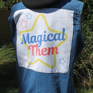 May include: A blue denim vest featuring a large white rectangular patch on the back. The patch displays a yellow star outline with the blue text "Magical" and the red text "Them" inside, surrounded by smaller red and blue star outlines, topped by a black beret.