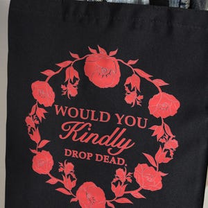 May include: Black tote bag with a red floral wreath design and the text "WOULD YOU KINDLY DROP DEAD." The bag has black handles and is made of a sturdy fabric. The design features stylized red roses.
