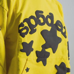 K&ouml;nnte beinhalten: Gelber Kapuzenpullover mit einem gro&szlig;en schwarzen Grafikdesign auf der Vorderseite. Das Design enth&auml;lt den Text "sp5der" in einer stilisierten Schriftart sowie unregelm&auml;&szlig;ige Formen und sternf&ouml;rmige Figuren. Der Hoodie hat eine Vordertasche.