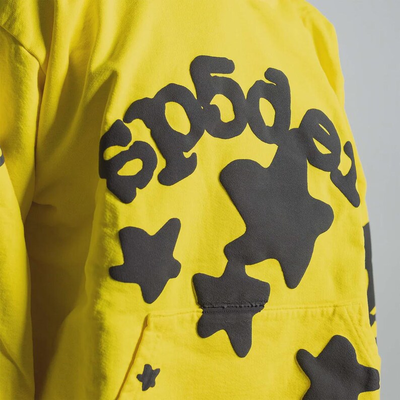 K&ouml;nnte beinhalten: Gelber Kapuzenpullover mit einem gro&szlig;en schwarzen Grafikdesign auf der Vorderseite. Das Design enth&auml;lt den Text "sp5der" in einer stilisierten Schriftart sowie unregelm&auml;&szlig;ige Formen und sternf&ouml;rmige Figuren. Der Hoodie hat eine Vordertasche.