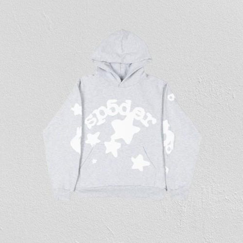 K&ouml;nnte beinhalten: Ein hellgrauer Kapuzenpullover mit Kapuze und K&auml;ngurutasche. Auf der Vorderseite steht das Wort "sp5der" in wei&szlig;er, stilisierter Schrift, zusammen mit wei&szlig;en Sternen. Die &Auml;rmel haben ebenfalls wei&szlig;e Grafiken. Der Hoodie ist aus weichem Material.