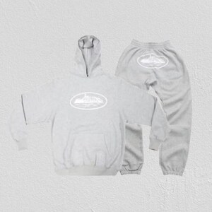 Op de afbeelding: Een lichtgrijze hoodie en joggingbroek set. De hoodie heeft een voorzak en een capuchon. Beide stukken hebben een wit ovaal ontwerp met een landschapsafbeelding. De joggingbroek heeft een elastische tailleband en manchetten.