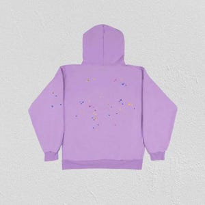 K&ouml;nnte beinhalten: Ein helllila Kapuzenpullover mit Kapuze und langen &Auml;rmeln. Auf der R&uuml;ckseite des Kapuzenpullovers befinden sich kleine, farbige Punkte in Gelb, Blau und Orange. Der Kapuzenpullover ist aus weichem Material.