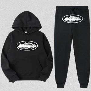 Op de afbeelding: Zwarte hoodie en joggingbroek set. De hoodie heeft een capuchon met trekkoord en een voorzak. Beide items tonen een witte ovale afbeelding met een gedetailleerde illustratie van een gebouw. De joggingbroek heeft een elastische tailleband en manchetten.