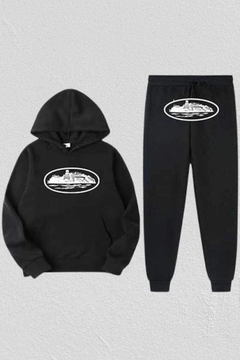 Op de afbeelding: Zwarte hoodie en joggingbroek set. De hoodie heeft een capuchon met trekkoord en een voorzak. Beide items tonen een witte ovale afbeelding met een gedetailleerde illustratie van een gebouw. De joggingbroek heeft een elastische tailleband en manchetten.