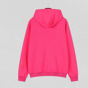 Puede incluir: Una sudadera con capucha rosa brillante con capucha a juego y pu&ntilde;os y dobladillo acanalados. Las mangas largas presentan peque&ntilde;as decoraciones doradas. La sudadera se muestra sobre un fondo liso de color claro.