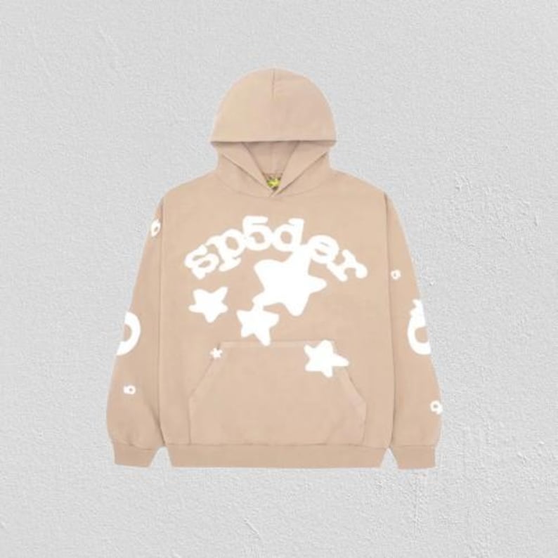 K&ouml;nnte beinhalten: Ein beigefarbener Hoodie mit einer Vordertasche und einer Kapuze. Der Hoodie zeigt den Text "sp5der" in Wei&szlig; sowie wei&szlig;e Sterngrafiken. Zus&auml;tzliche wei&szlig;e Grafiken befinden sich auf den &Auml;rmeln. Der Hoodie besteht aus einem weichen Material.