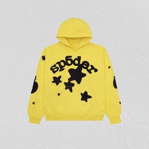 K&ouml;nnte beinhalten: Ein gelber Kapuzenpullover mit dem Wort "sp5der" in Schwarz sowie schwarzen Sternen. Der Pullover hat eine K&auml;ngurutasche und schwarze kreisf&ouml;rmige Designs auf den &Auml;rmeln.