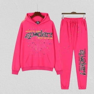 Puede incluir: Un conjunto de ch&aacute;ndal rosa brillante que consta de una sudadera con capucha y pantalones a juego. Tanto la sudadera con capucha como los pantalones muestran la palabra "SPYDER" en una fuente estilizada. Un conjunto llamativo.
