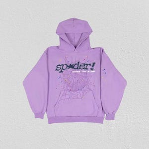 K&ouml;nnte beinhalten: Ein lila Kapuzenpullover mit einem Spinnennetz-Design und dem Wort "spider!" in einer pixeligen Schriftart. Der Hoodie hat eine K&auml;ngurutasche und eine Kapuze mit Kordelzug. Die Grafik enth&auml;lt bunte Punkte.