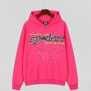 Puede incluir: Una sudadera con capucha de color rosa brillante con un bolsillo delantero y una capucha. La parte delantera presenta la palabra "Spider" en una fuente estilizada, con un dise&ntilde;o de telara&ntilde;a y el texto "defended the punk" y "bares the alibi".