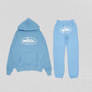 Op de afbeelding: Een lichtblauwe hoodie en bijpassende joggingbroek set. Beide items hebben een witte ovale afbeelding met een gedetailleerde illustratie. De hoodie heeft een voorzak en een capuchon. De joggingbroek heeft een elastische tailleband.