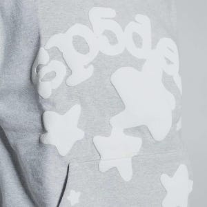K&ouml;nnte beinhalten: Nahaufnahme eines hellgrauen Hoodies mit wei&szlig;en Stern- und Textapplikationen. Das Wort "good" ist in einer stilisierten Schriftart zu sehen. Der Hoodie hat eine K&auml;ngurutasche und eine Kapuze mit Kordelzug.
