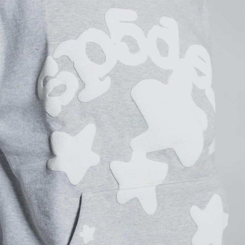 K&ouml;nnte beinhalten: Nahaufnahme eines hellgrauen Hoodies mit wei&szlig;en Stern- und Textapplikationen. Das Wort "good" ist in einer stilisierten Schriftart zu sehen. Der Hoodie hat eine K&auml;ngurutasche und eine Kapuze mit Kordelzug.
