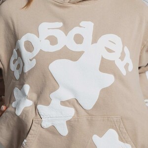 K&ouml;nnte beinhalten: Beigefarbener Kapuzenpullover mit wei&szlig;em Grafikdesign. Das Design beinhaltet den Text "5dge" in einer stilisierten Schriftart sowie sternf&ouml;rmige und abstrakte Formen. Der Kapuzenpullover hat eine K&auml;ngurutasche und eine Kapuze mit Kordelzug.