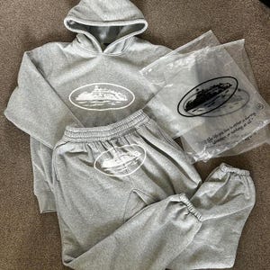 Op de afbeelding: Een grijze hoodie en bijpassende joggingbroek set. De hoodie heeft een capuchon met trekkoord en een grote witte ovale afbeelding op de voorkant. De joggingbroek heeft een elastische tailleband en manchetten, met een bijpassende afbeelding.