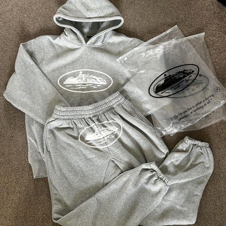 Op de afbeelding: Een grijze hoodie en bijpassende joggingbroek set. De hoodie heeft een capuchon met trekkoord en een grote witte ovale afbeelding op de voorkant. De joggingbroek heeft een elastische tailleband en manchetten, met een bijpassende afbeelding.